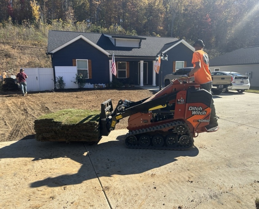 Mini Skid Man in orange shirt operates a mini skid steer in front of a single story blue house. He is carrying a pallet of sod